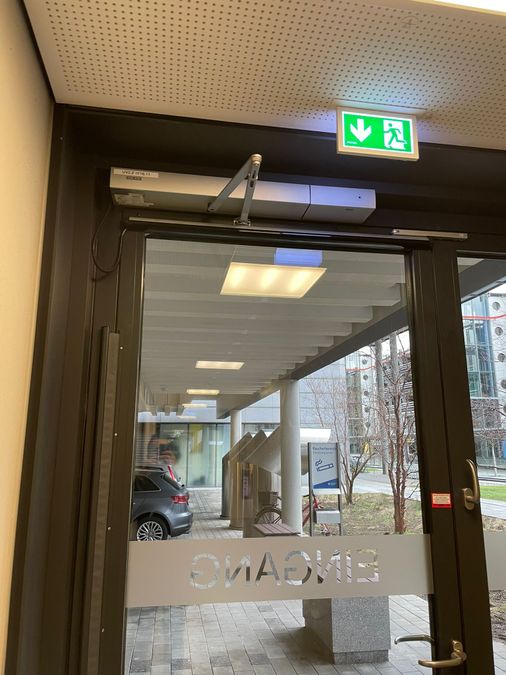 Gläserne Eingangstür mit obenliegendem Antrieb und Fluchtwegkennzeichnung