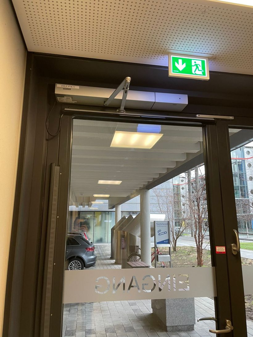 Gläserne Eingangstür mit obenliegendem Antrieb und Fluchtwegkennzeichnung